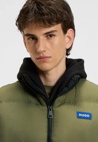 Chaqueta verde oliva con capucha negra, cremallera frontal y parche de logo azul. El material parece acolchado con una textura suave.