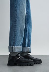 Black leather shoes with a chunky sole and decorative buckle detail. Worn with cuffed blue jeans featuring a lighter cuff.