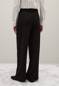 Zwart wijde broek gemaakt van een gladde stof, met riemlussen en twee achterzakken. Gepaard met zwart versierde schoenen.