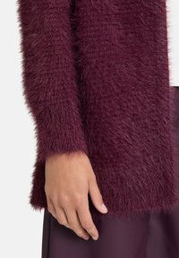 Dłoń spoczywa obok burgundowego, puszystego swetra z długim rękawem oraz pasującej spódnicy lub spodni, z częściowo widoczną białą odzieżą.