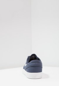Sneaker in tela blu navy con suola bianca e logo Nike, visto da dietro su una superficie bianca con uno sfondo di angolo bianco semplice.
