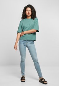 Türkiissinist värvi üle mõõtude t-särk, mis on valmistatud puuvillast, kombineeritud helesiniste kitsaste teksapükstega ja mustade sandaalidega, esitletud neutraalsel taustal.