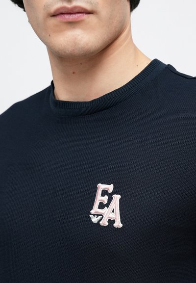 Μπλε ναυτικό πλεκτό πουλόβερ με υφή, με ροζ κεντητό λογότυπο "EA" στο στήθος. Στρογγυλή λαιμόκοψη με ριμπ τελείωμα.