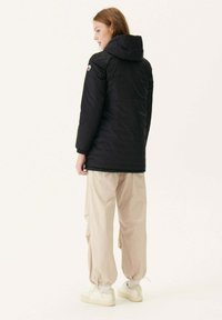Giacca a vento nera con cappuccio, polsini elasticizzati e patch con logo. Abbinata a pantaloni beige a sigaretta e sneakers chiare. Design minimalista.
