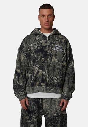 Mężczyzna w czarno-szarej bluzie na zamek z wzorem i pasujących spodniach, z białym napisem na bluzie: "Good for Nothing British Atelier."