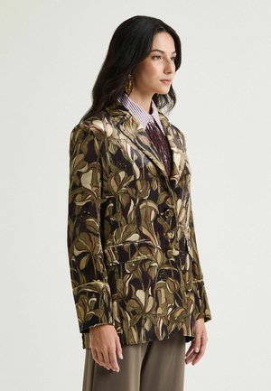 Femme aux cheveux foncés porte un blazer à motifs aux tons terre sur une chemise rayée et un pantalon de couleur neutre, debout devant un fond uni.