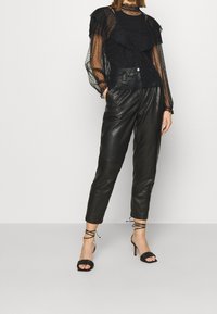 Schwarze, mehrlagige Bluse mit transparenten Puffärmeln, kombiniert mit schwarzen, taillierten Lederhosen und sandalenartigen, hohen Absätzen.