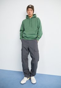 Sweat à capuche vert avec poche avant, pantalon cargo gris avec poches latérales, et baskets blanches avec accents jaunes. Le modèle se tient devant un fond clair.