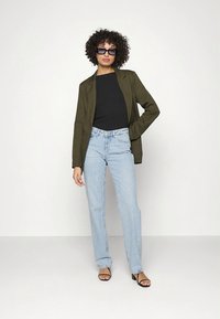 Χακί πράσινο σακάκι πάνω από μαύρο τοπ, σε συνδυασμό με ανοιχτό μπλε τζιν ψηλόμεσα και μαύρα πέδιλα με τακούνι· μοντέλο με κατσαρά μαλλιά και γυαλιά ηλίου.