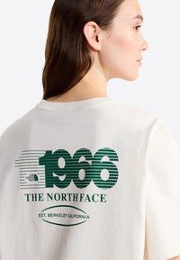 Mujer de cabello castaño con una camiseta blanca que lleva texto verde "1966 The North Face Est. Berkeley California" en la espalda, mirando hacia la derecha.