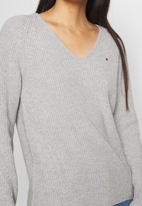 Pull tricot gris clair avec col en V, motif texturé et manches longues. Petit logo distinctif sur la poitrine.