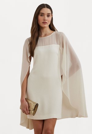 Robe courte crème avec des manches en cape transparentes, un col rond et une texture lisse. Accompagnée d'un sac à main doré avec une finition à motifs.