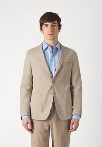 Blazer beige in cotone con due tasche frontali, chiusura a un bottone, rever a punta; abbinato a una camicia azzurro chiaro con colletto.