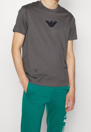 Homme portant un t-shirt gris foncé à manches courtes avec un emblème noir sur la poitrine et un pantalon turquoise avec un texte blanc imprimé sur la jambe gauche.