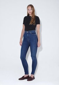 Jeans azul de cintura alta con un diseño ajustado, que presentan una textura suave y costuras oscuras, combinados con una blusa negra de manga corta y mocasines marrones.