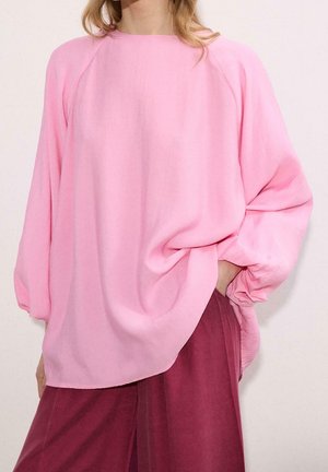 Femme portant un chemisier rose ample à manches longues et un pantalon bordeaux taille haute, une main dans la poche.