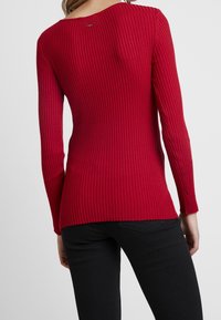Rood geribd longsleeve top met een brede ronde hals, met een subtiele textuur en gespleten zoom; achterkant heeft een klein logo detail. Gecombineerd met zwarte broek.