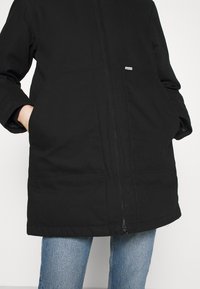 Sort jakke med lynlås og en afslappet pasform, to forlommer og et lille mærke. Bruges over lyseblå jeans med en glat stoftekstur.