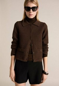 Cardigan tricotat texturat, de culoare maro, cu guler, închidere cu nasturi și două buzunare. Purtați-l peste un top maro, cu șorturi negre și ochelari de soare.