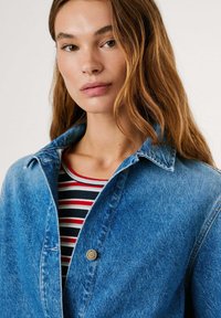 Blauwe denimjas met een kraag, voorzien van een gouden knoopdetail. Eronder een gestreept shirt met rood, wit en marineblauw.