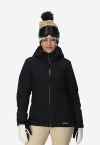 Giacca da sci impermeabile nera con chiusura a zip, cappuccio regolabile e numerose tasche. Indossata con guanti e un cappello beige con pon pon.