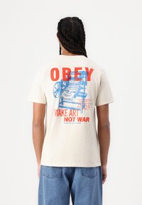 Beige cotton t-shirt featuring bold red and blue graphic text on the back: "OBEY MAKE ART NOT WAR," with a design of a printing press.