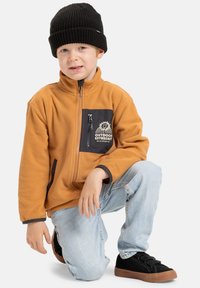 Fleeceová bunda oranžové barvy s černou zipovou kapsou, která obsahuje tištěný design. Černá pletená čepice a modré džíny doplňují černé tenisky.