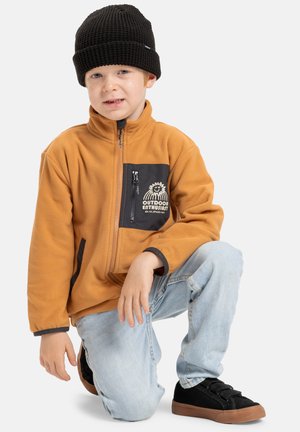 Veste en polaire orange avec poche zippée noire, ornée d'un design imprimé. Bonnet en tricot noir et jean bleu complètent des baskets noires.