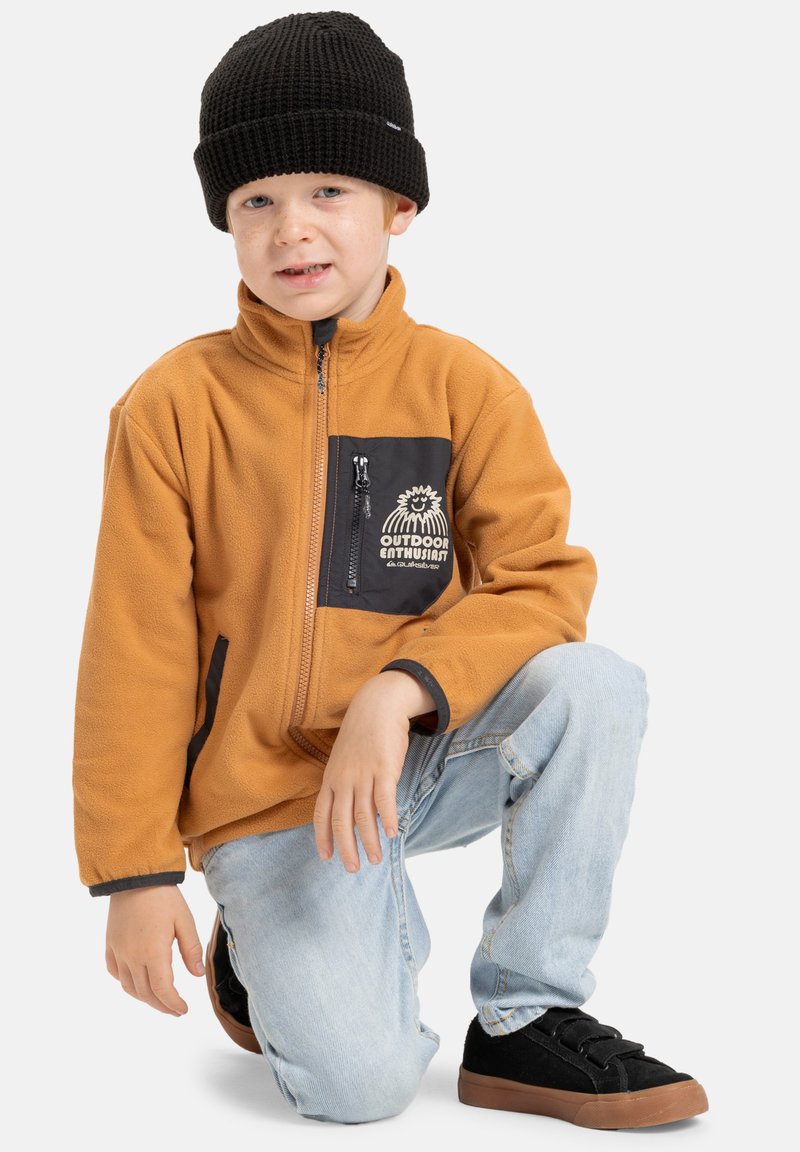 Fleeceová bunda oranžové barvy s černou zipovou kapsou, která obsahuje tištěný design. Černá pletená čepice a modré džíny doplňují černé tenisky.