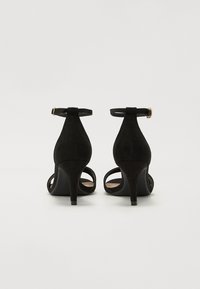 Sandalias de tacón alto de ante negro con correas en el tobillo, punta abierta y tacones cónicos. El detalle de hebilla dorada en las correas realza el diseño.
