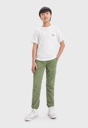 T-shirt bianco in cotone con taschino sul petto e pantaloni cargo verdi con multiple tasche, polsini affusolati e sneakers bianche con dettagli rossi.