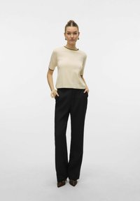 Modell iklädd en ljus krämfärgad topp med svart kant och svarta högmidjade byxor, händerna i fickorna, med en minimalistisk bakgrund.