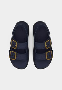 Ζευγάρι σκούρα μπλε σανδάλια με δύο ρυθμιζόμενους υφασμάτινους ιμάντες και πόρπες σε χρυσό τόνο, απεικονιζόμενα από πάνω σε λευκό φόντο.