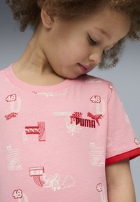 Gaiši rozā T-kreklā ar sarkaniem akcentiem, kas izceļas ar izsmeļošām grafikām un tekstu, tostarp Puma logotipu, dažādās krāsās un dizainos.