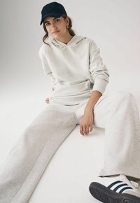 Hellgrauer Kapuzenpullover, passende Jogginghose und schwarze Mütze. Sneaker mit schwarz-weißen Streifen. Weicher Stoff.
