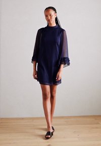 Navy blue dress with sheer, ruffled bell sleeves, a high neckline, and a relaxed fit. The model wears black heeled slides. Light wood floor.