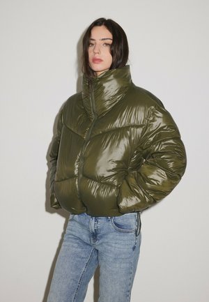 Femme aux cheveux foncés portant une doudoune vert olive brillante et un jean bleu clair, debout les mains dans les poches devant un fond uni.