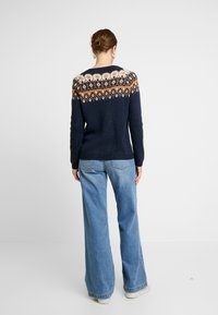 Pull en tricot bleu marine avec un empiècement à motif comprenant des losanges blancs, orange et marron ; associé à un jean bleu clair à jambes larges.