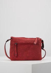 Sac bandoulière perforé rouge avec poche zippée à l'avant et bandoulière réglable, exposé sur une surface blanche avec un fond clair uni.
