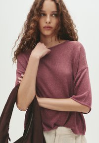 Malvino strukturiertes Strickoberteil in tiefem Mauve mit einem weiten Ausschnitt und kurzen Ärmeln. Kombiniert mit einer leichten braunen Jacke, die über dem Arm getragen wird.