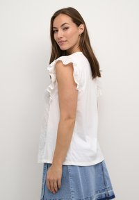 Camisa de algodón blanca con mangas con volantes y borde de ojal. La parte posterior muestra una textura suave, combinada con una falda de mezclilla.