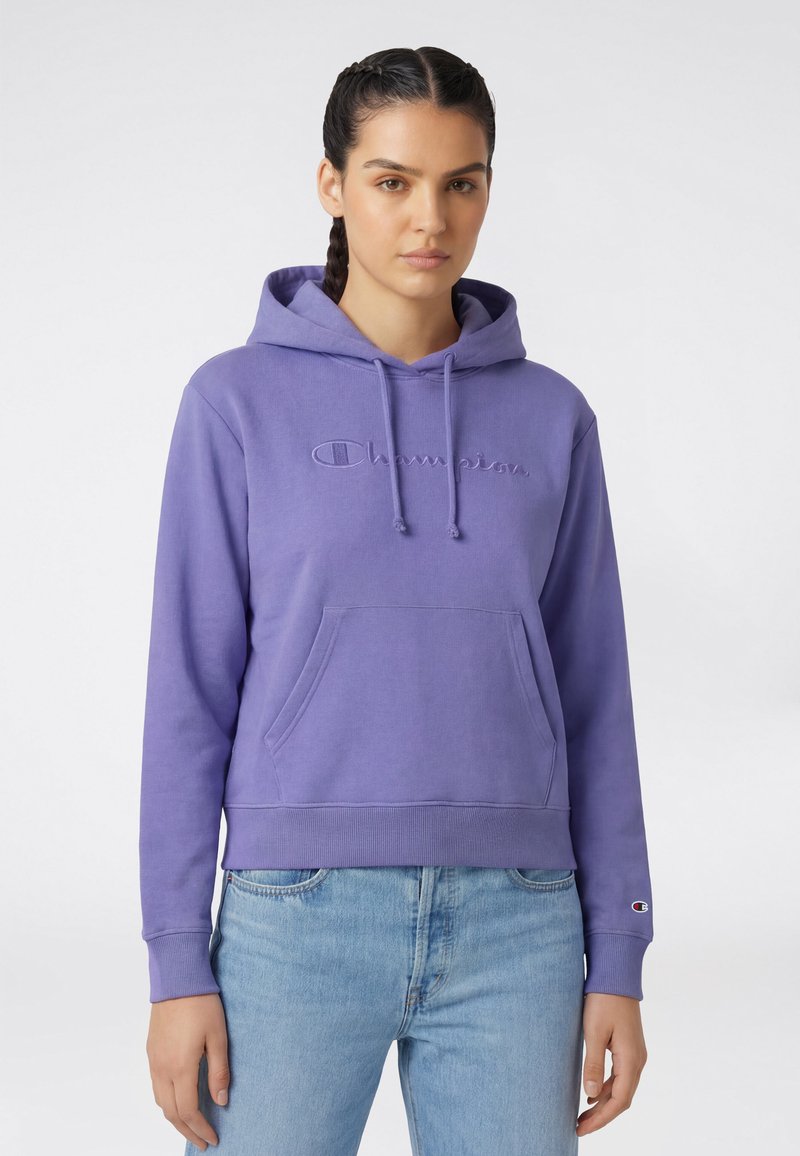 Frau mit geflochtenem Haar, die einen lila Champion-Hoodie mit Fronttasche und hellblaue Jeans trägt, stehend vor einem einfarbigen Hintergrund.