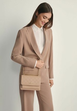 Kobieta w beżowym garniturze trzymająca dopasowaną beżową torebkę z złotym łańcuszkiem, uśmiechająca się i spoglądająca w dół na jednolitym tle.