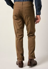 Hnědé bavlněné kalhoty s přiléhavým střihem, s ohrnutými nohavicemi, dvěma zadními kapsami a jemnými švy. Nosené s hnědými koženými botami.