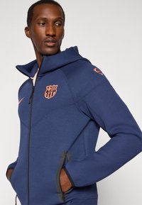 Homme portant un sweat à capuche zippé bleu marine avec des logos orange de FC Barcelone et Nike, les mains dans les poches, regardant à sa gauche sur un fond uni.