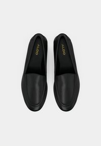 Chaussures noires à enfiler en cuir lisse, avec un bout arrondi, présentant des coutures minimalistes et un logo doré sur la semelle intérieure.
