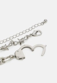Metal handcuff accessory with a shiny silver finish, linked by a chain with a clasp. Features detailed cutouts and a textured grip.