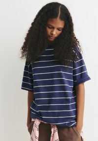 Marineblå stribede t-shirt med korte ærmer, løs pasform og hvide vandrette striber. Sættes sammen med brune bukser og et lyserødt stribet slips.