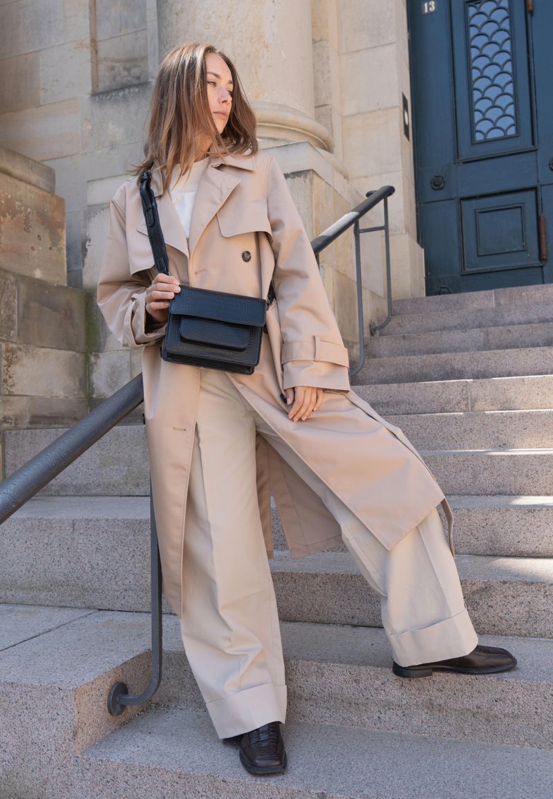 Vrouw die een beige trenchcoat en wijde broek draagt, een zwarte crossbodytas vasthoudend, staand op stenen treden naast een donkerblauwe deur.