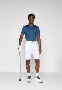 Dark blue polo shirt with a geometric pattern, white shorts, white shoes with blue accents, holding a golf club, wearing a white glove.
